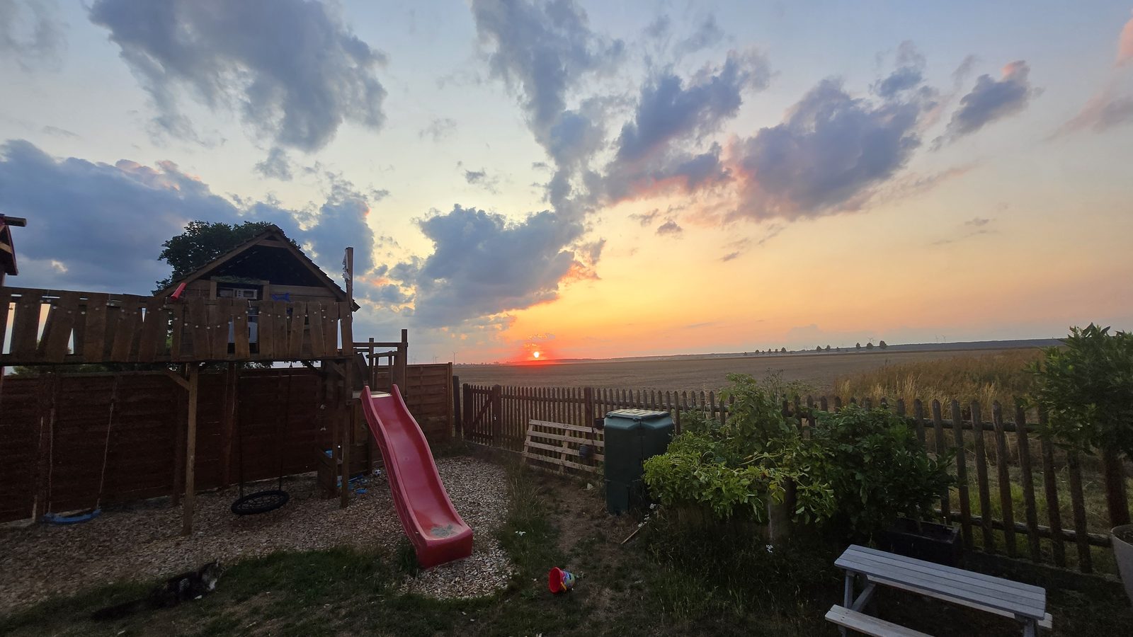 Garten mit Spielturm, Rutsche und Schaukel – dahinter freier Blick über die Felder in den Sonnenuntergang